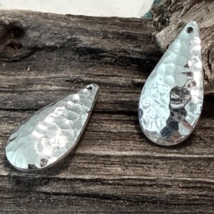 Può includere: Due ciondoli in metallo color argento a forma di goccia. Ogni ciondolo presenta una superficie martellata e testurizzata che riflette la luce e include un piccolo foro circolare nella parte superiore per l'aggancio, creando un aspetto metallico.