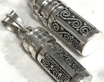 May include: Two silver-colored pendant necklaces. Each pendant is cylindrical with ornate, detailed scrollwork and a screw-top design. The pendants are suspended from a silver bail. The background is a light, neutral color.