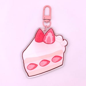 May include: A pink and white acrylic keychain shaped like a slice of strawberry shortcake. The keychain features a pink and white frosting design with three red strawberries on top.