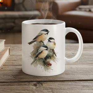 Puede incluir: Taza de café de cerámica blanca con una pintura de acuarela de tres pequeños pájaros posados en una rama con una piña. Los pájaros tienen cabezas negras y blancas y cuerpos de color canela. Sale vapor del líquido oscuro dentro de la taza.