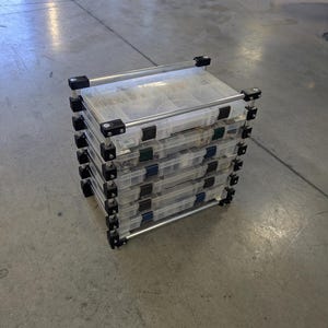 May include: A stack of clear plastic storage boxes with black and silver metal supports. Each box has a hinged lid and is filled with various small items. The boxes are stacked on top of each other on a concrete floor.