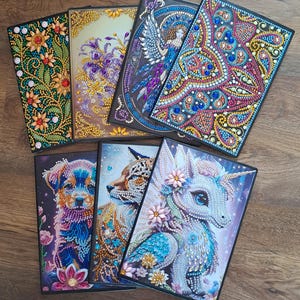 May include: A selection of decorative notebooks with detailed, vibrant designs. The notebooks showcase images of flowers, a dog, a fox, a unicorn, and a winged woman. Each notebook is adorned with sparkling embellishments.