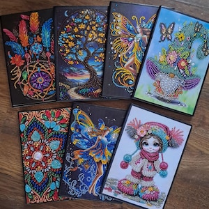 May include: A selection of seven diamond painting kits with diverse designs. The designs include a dreamcatcher, a tree, a fairy, a gnome, a mandala, a fairy, and a girl in winter attire. Each kit has a black border.