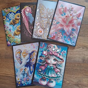 May include: Six framed diamond art paintings are displayed. The artwork includes bees, a bird, a butterfly, flowers, an elephant, and a doll. The paintings are vibrant and detailed, with a sparkling, textured finish, adding a touch of elegance.