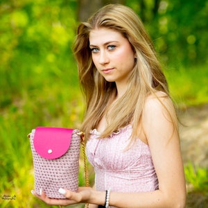 May include: A young woman with long blonde hair is holding a pink crocheted bag with a pink leather flap and a gold chain strap. The bag has a small white button on the flap.