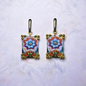 May include: A pair of gold earrings with a rectangular floral design. The earrings feature a blue flower with red and white accents, surrounded by a gold frame with small gold beads.