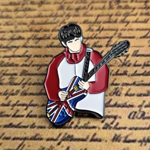 Peut inclure: Épingle en émail représentant une figurine jouant de la guitare avec un motif Union Jack. La figurine porte une tenue rouge et blanche. L'épingle est posée sur un fond de texte manuscrit.