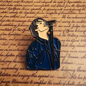 Peut inclure: Une épingle en émail noir et bleu représentant une personne chantant dans un microphone. La personne porte une veste bleue et a les cheveux foncés.