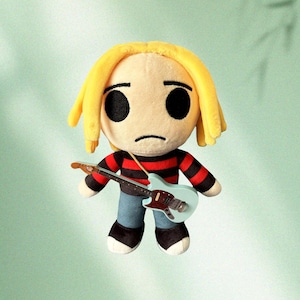 May include: A plush doll of a musician with yellow hair, black eyes, and a sad expression. The doll wears a red and black striped shirt, blue jeans, and black shoes. It holds a light blue guitar with a brown neck.