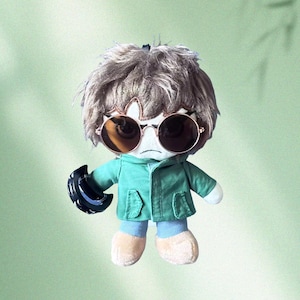 Puede incluir: Muñeco de peluche con pelo castaño, gafas de sol redondas y una chaqueta verde. El muñeco sostiene un objeto negro y tiene una expresión neutra. El fondo es verde claro.