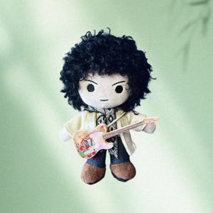 May include: A plush doll of a musician with black curly hair, wearing a cream-colored jacket, blue jeans, and brown shoes. The doll holds a colorful guitar with a pink and white body. The doll has a neutral expression.