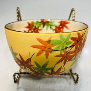 Puede incluir: Un cuenco amarillo con borde dorado y soporte decorativo. El cuenco está adornado con un patrón de hojas de arce naranjas, verdes y marrones. El soporte es de metal dorado.