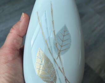 May include: White ceramic vase with a teardrop shape. The vase features a gold and gray leaf and bamboo design. The vase is held in a hand.