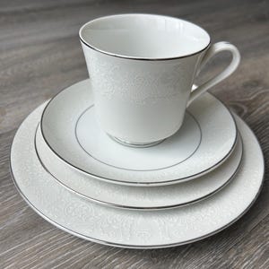 May include: A white porcelain teacup and saucer set with a floral design and silver edging. The set includes a teacup, a saucer, and two plates. The teacup sits on the saucer, with the plates stacked beneath. The plates and cup have a delicate pattern.