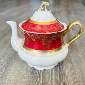 May include: A white and red porcelain teapot with gold accents. The teapot features a red band with gold floral designs and gold trim. The handle and spout are white, and the lid is white with a gold finial.