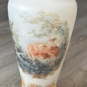 May include: White porcelain vase with a gold rim and a painted scene of a woman on a swing in a garden. The vase has a flared base and a narrow neck. Floral designs decorate the bottom of the vase.