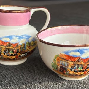 May include: A vintage cream jug and bowl set. The jug and bowl are white with a pink rim and gold trim. The set features a colourful illustration of a town scene with a horse-drawn carriage.