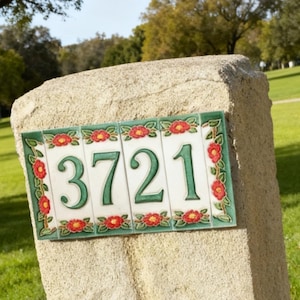 Pode incluir: Placa de endereço retangular com o número 3721 em verde sobre fundo branco. A placa é emoldurada com uma borda verde e detalhes florais vermelhos. Montada em um pilar de pedra.