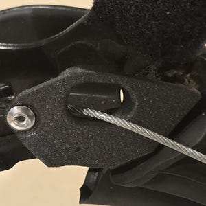 May include: Close-up of a black mechanical component with a silver cable running through it. The component has a textured surface and a metal fastener. The image highlights the intricate design and construction of the part.