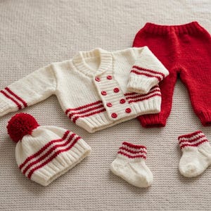May include: A knitted baby outfit in cream and red. The set includes a cardigan with red buttons and stripes, matching pants, a hat with a pom-pom, and two pairs of socks. The outfit is made of soft yarn.