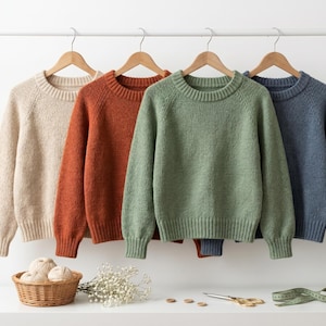 Op de afbeelding: Vier crewneck truien in beige, roest, groen en blauw hangen aan houten hangers. Een klein mandje met garen, bloemen, een schaar en een meetlint staan op het witte oppervlak eronder. De truien hebben geribde manchetten en kragen.