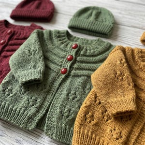 May include: Close-up of three knitted baby cardigans in burgundy, green, and mustard yellow, each with matching beanies. The cardigans have red ladybug-shaped buttons and intricate knit patterns. The items are arranged on a light wooden surface.