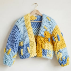 May include: A chunky knit cardigan in shades of light blue and yellow, with decorative yellow and blue stitching. The open-front sweater is displayed on a wooden hanger against a white background. A cozy, handmade garment.
