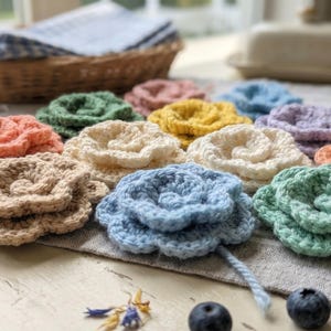 Könnte beinhalten: Eine Sammlung mehrfarbiger gehäkelter Blumen in Blau-, Grün-, Gelb-, Rosa-, Beige- und Weißtönen. Jede Blume weist ein geschichtetes Spiralblütenblatt-Design aus Garn auf, die in einer Gruppe auf einer hellen Stoffoberfläche angeordnet sind.