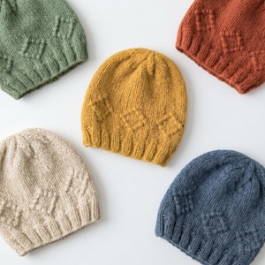 May include: Five knitted beanies in various colors: green, mustard yellow, rust, beige, and blue. Each beanie features a diamond pattern. The beanies are displayed on a white surface.