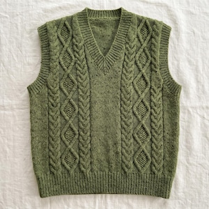 May include: A green knitted vest with a V-neck. The vest features a cable knit design with diamond and braid patterns. The sleeveless garment is made of a textured, speckled yarn. The vest is displayed on a white surface.