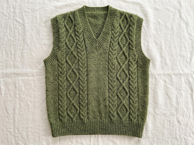 May include: A green knitted vest with a V-neck. The vest features a cable knit design with diamond and braid patterns. The sleeveless garment is made of a textured, speckled yarn. The vest is displayed on a white surface.