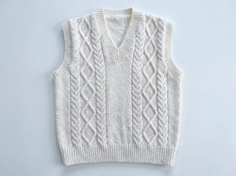 May include: Cream-colored cable knit sweater vest with a V-neck. The sleeveless vest features a textured cable knit design on the front and back. The ribbed hem and neckline add a classic touch to this cozy garment.