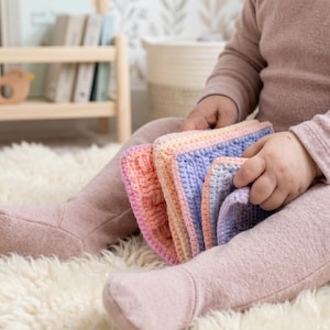 Puede incluir: Una pila de cuadrados de ganchillo hechos a mano en tonos rosa, naranja y azul. Los cuadrados son sostenidos por una persona que lleva un atuendo rosa, sentada en una alfombra blanca y esponjosa. Una estantería de madera y una decoración con forma de pájaro están en el fondo.