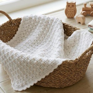 Può includere: Un cesto di vimini con una coperta bianca all'uncinetto. Il cesto ha due manici ed è riempito con una coperta morbida e strutturata. Giocattoli di legno e un uncinetto sono visibili.