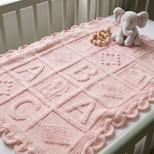 May include: A pale pink knitted baby blanket featuring the letters A, B, and C, and other shapes. The blanket has a scalloped edge and is on a cot. A plush elephant and wooden toy are on the blanket.