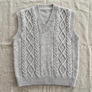 May include: A light gray cable-knit sweater vest with a V-neck. The vest features a textured cable-knit design on the front and back panels, with ribbed detailing at the hem and neckline. The sleeveless garment is made of a soft, speckled yarn.