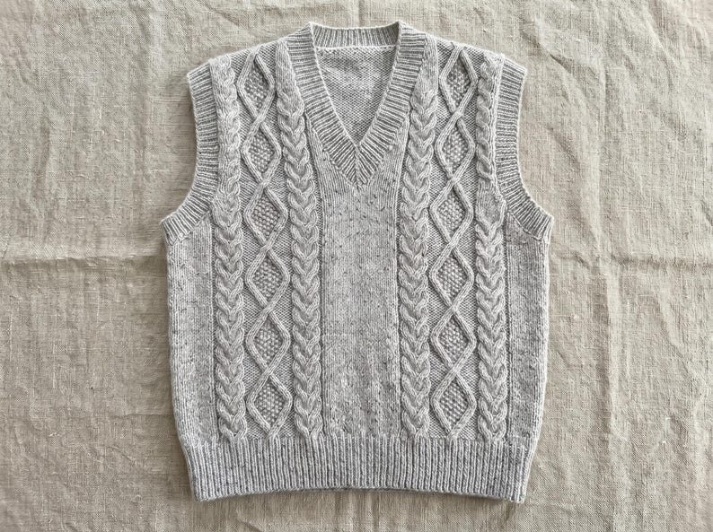 May include: A light gray cable-knit sweater vest with a V-neck. The vest features a textured cable-knit design on the front and back panels, with ribbed detailing at the hem and neckline. The sleeveless garment is made of a soft, speckled yarn.