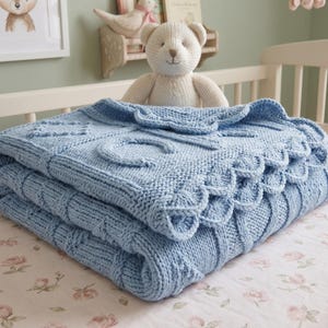 May include: A folded, light blue knitted baby blanket with a scalloped edge and raised letter details. The blanket is stacked on a floral patterned surface, with a crib and stuffed animals in the background.