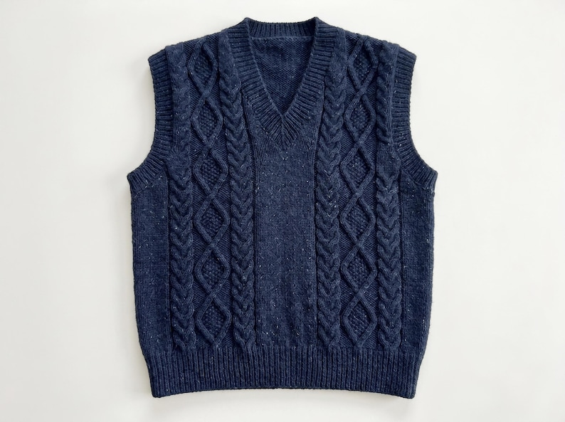 May include: A navy blue cable-knit sweater vest with a V-neck. The vest features a textured pattern with vertical cable designs and a ribbed hem. The sleeveless garment is made of a soft, speckled yarn.