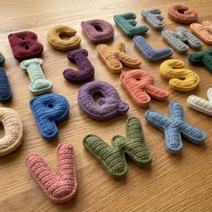 Puede incluir: Un conjunto de letras del alfabeto de ganchillo, coloridas, dispuestas sobre una superficie de madera. Las letras son de varios colores, incluyendo verde, azul, rosa y amarillo. Las letras son suaves y texturizadas, ideales para fines educativos.