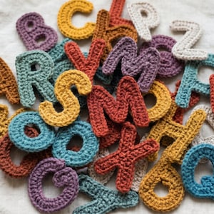 May include: A pile of colourful, crocheted letters in shades of orange, yellow, blue, green, purple, and brown. The letters are scattered, showing the handmade texture and detail of each piece. The letters are likely for crafting or educational use.