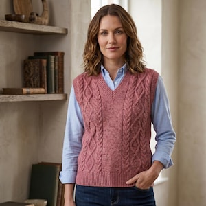 May include: A woman wearing a dusty rose cable-knit sweater vest over a light blue button-down shirt and blue jeans. The vest has a V-neck and intricate cable patterns. The background includes a bookshelf and a window.