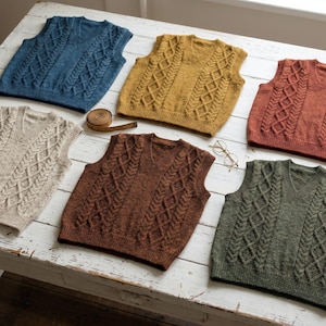 May include: Six cable-knit vests in various colors, including blue, mustard yellow, and rust, are arranged on a white wooden surface. Each vest features a V-neck and a central cable design. A measuring tape and eyeglasses are also present.