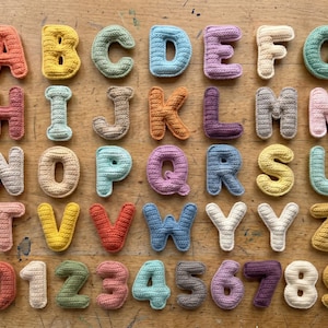 Könnte beinhalten: Eine Sammlung farbenfroher, gehäkelter Buchstaben und Zahlen, die auf einer Holzoberfläche angeordnet sind. Die Buchstaben sind in verschiedenen Farben gehalten, darunter Orange, Gelb, Blau und Lila. Die Zahlen 0 bis 9 sind ebenfalls enthalten, in einer Vielzahl von Farben.