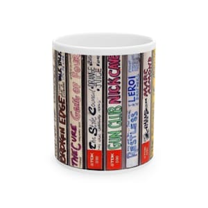 May include: White ceramic mug featuring a design of vintage cassette tapes. The tapes display band names like "The Cure," "Nick Cave," and "Marc Almond." The mug is a standard size and shape, perfect for coffee or tea.
