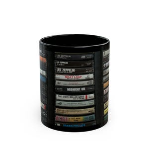 May include: Black ceramic mug with a design of stacked cassette tapes featuring various artists and album titles, including Led Zeppelin, Meat Loaf, Midnight Oil, The Steve Miller Band, Tom Petty, and Shawn Phillips.