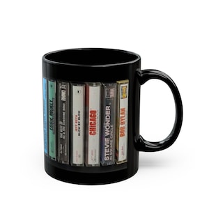 May include: Black ceramic coffee mug featuring a row of vintage cassette tapes. The tapes include artists like Eddie Money, KC & The Sunshine Band, Jeff Beck, Chicago, Stevie Wonder, and Bob Dylan. A music-themed gift.