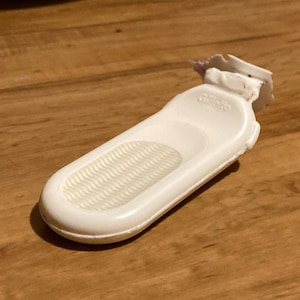 May include: A white Gillette razor with a textured grip and the brand name printed on the handle. The razor head is white with a small portion of the blade visible. The razor is on a wooden surface.