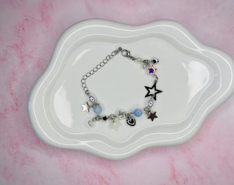 May include: A silver-toned charm bracelet with star, heart, and bead charms. The bracelet features a variety of charms, including a black star, a silver star, a heart, and light blue beads. The bracelet is displayed on a white, cloud-shaped plate.
