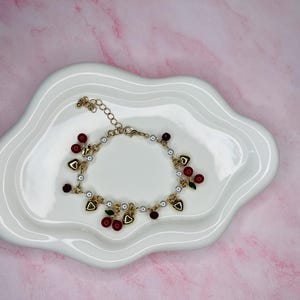 May include: A gold-toned charm bracelet with red cherry and heart-shaped charms, white beads, and small gold accents. The bracelet is displayed on a white, wavy-edged plate against a pink marble background.